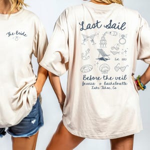 May include: Beige t-shirts with navy blue text and graphics. One shirt says "The Bride" with a ring graphic. The other says "Last Sail" with beach-themed illustrations and "Before the veil" with a location.