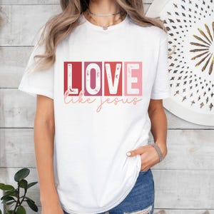 Puede incluir: Camiseta blanca con la palabra "LOVE" en un diseño de letras en bloque, cada letra en un tono diferente de rojo y rosa. Debajo de la palabra "LOVE" está la frase "like jesus" en una fuente cursiva. La camiseta es de estilo casual.
