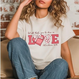 Puede incluir: Camiseta color crema con un corazón rosa y la palabra "LOVE" en letras rosas. El corazón está decorado con pequeños corazones rojos y un lazo rosa. El texto "Let all that you be done in" y "1 Corintios 16:14" también están impresos en la camiseta.