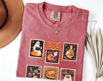 May include: A watermelon-colored t-shirt featuring Halloween-themed postage stamp designs. The stamps depict ghosts, pumpkins, and a cake, with the word "BOO!" on one. A silver necklace and a brown hat are also visible.