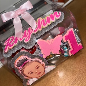 Puede incluir: Una caja de regalo de plástico transparente atada con un lazo de satén rosa. La caja está decorada con detalles de purpurina rosa y plateada, una mariposa rosa y el número 1. La caja también presenta la cara de un bebé y el nombre "Rhythm" en rosa.