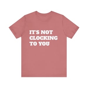 May include: A dusty rose t-shirt with the white text "IT'S NOT CLOCKING TO YOU" printed on the front. The shirt has short sleeves and a crew neck. The fabric appears to be soft and comfortable.