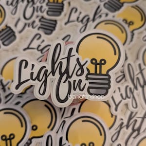 May include: A sticker with the words "Lights On" in black script, featuring a yellow lightbulb design. The sticker has a white border and the shop name "banatenshop" is at the bottom. Other similar stickers are in the background.