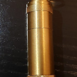以下が含まれることがあります： 上部に小さなリングが付いた金色をした金属製の容器。容器の上部と下部には、テクスチャ加工が施されています。