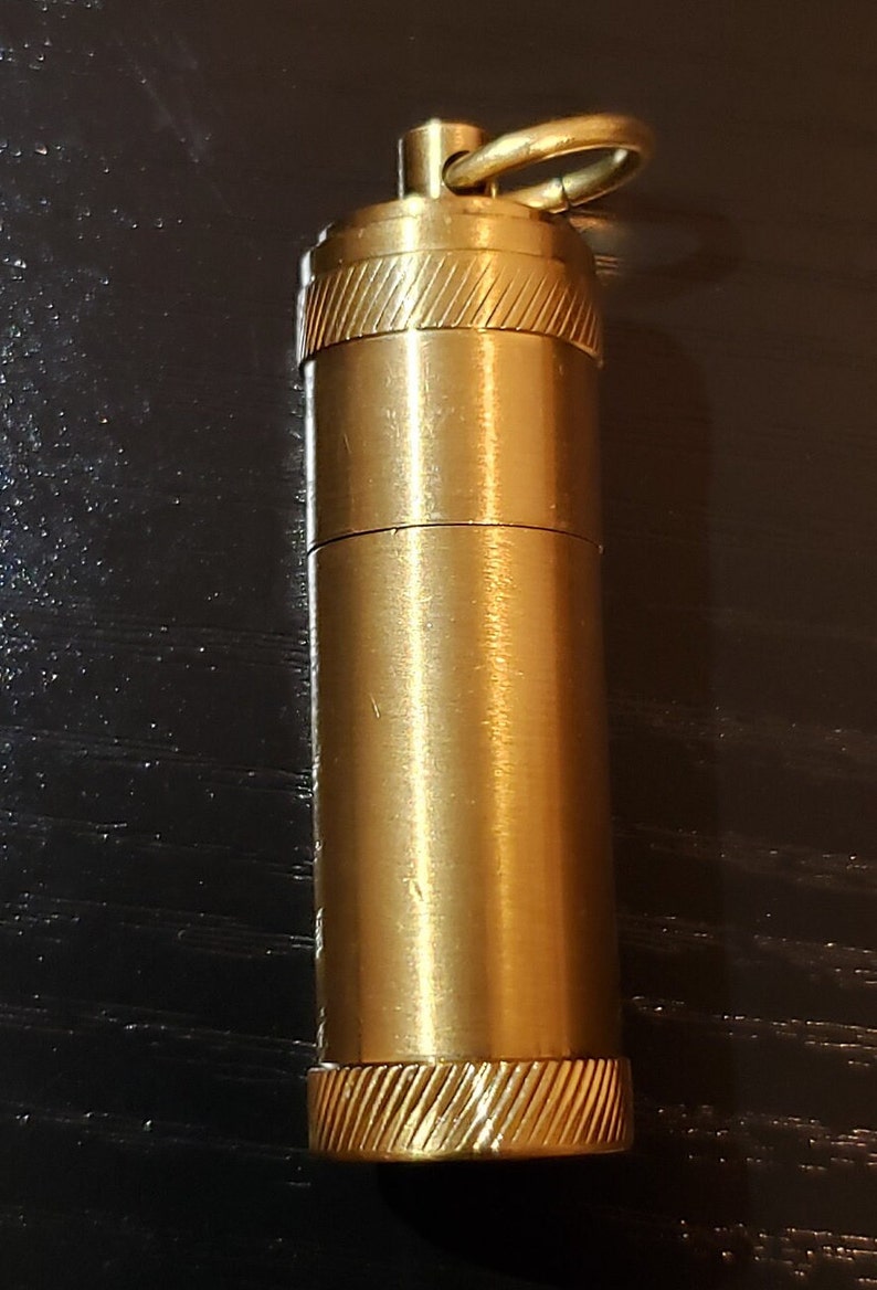 以下が含まれることがあります： 上部に小さなリングが付いた金色をした金属製の容器。容器の上部と下部には、テクスチャ加工が施されています。