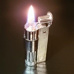 Puede incluir: Un encendedor plateado con una llama que sale de la parte superior. El encendedor tiene un diseño texturizado y una pequeña palanca en el lateral.