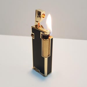 May include: A black and gold lighter with a flame coming out of the top. The lighter has a gold trim and a black enamel finish.