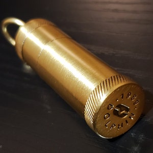 以下が含まれることがあります： 上部にループが付いた金色の金属製容器。容器にはテクスチャパターンが施され、底面には「DOLPHIN 1788」という文字が刻まれています。