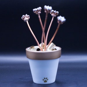 May include: A white ceramic flower pot with a gold rim and a paw print design. The pot is filled with spent bullet casings, and five bullet casings are arranged to look like flowers.