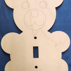 May include: A wooden light switch cover shaped like a bear. The bear has a simple face with two eyes, a nose, and a mouth. The switch cover is unfinished and ready to be painted.