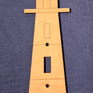 May include: A wooden lighthouse-shaped light switch cover with a light brown finish. The cover has a rectangular opening for the switch and four small circular holes.
