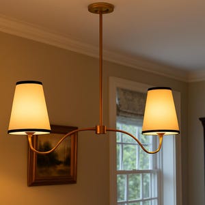 May include: A gold-toned, two-light pendant lamp with off-white shades and black trim. The lamp hangs from a ceiling mount and features a curved arm design. The shades are illuminated, casting a warm glow.