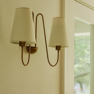 Puede incluir: Aplique de pared de dos luces con acabado de latón. La lámpara tiene dos pantallas cónicas de color blanquecino. El accesorio está montado en una pared de color claro, con una ventana visible en el fondo.