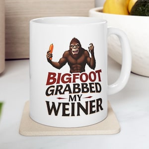 May include: White ceramic mug featuring a cartoon Bigfoot character holding a hot dog. The mug has the text "BIGFOOT GRABBED MY WEINER" in a bold, humorous font. The mug is on a beige coaster.