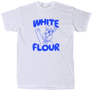 May include: White t-shirt with a blue graphic of a chef character and the words "WHITE FLOUR". The chef is wearing a toque and a neckerchief, with one arm raised in a gesture of triumph. The design is centered on the front of the shirt.