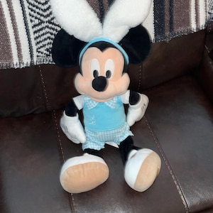 May include: A Mickey Mouse plush toy dressed in a light blue outfit with a bow tie and bunny ears. The plush has black ears, a blue headband, and white gloves and feet. The toy is sitting on a brown leather couch.