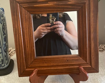May include: A brown wooden picture frame with a mirror inside. The frame is standing on a small wooden easel. The mirror is reflecting a person holding a phone.