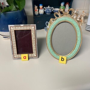 May include: Two decorative picture frames. One is rectangular with a silver and gold border, and the other is oval with a gold and teal frame. Both frames are empty and ready for photos.