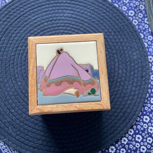 May include: Square wooden box with a decorative tile top. The tile features a Native American figure in a pink garment, with blue and green accents. The box has a light wood frame and is set against a blue background.