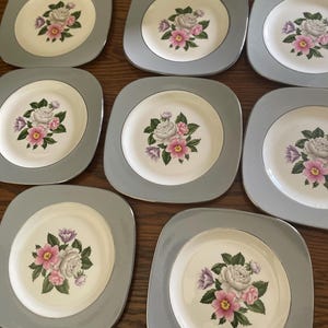 May include: Nine square-shaped plates with a gray border and a floral design. The center of each plate is off-white, featuring a bouquet of pink, white, and purple flowers with green leaves. The plates are arranged on a wooden surface.