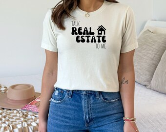 May include: A white t-shirt with black text that reads "Talk Real Estate To Me" in a retro font. The text is surrounded by a black house icon.