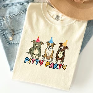 Pode incluir: Camiseta creme com um gráfico de três cães pit bull de desenho animado usando chapéus de festa. O texto "Pitty Party" é impresso em letras coloridas abaixo dos cães. A camisa está dobrada sobre uma superfície de jeans azul claro.