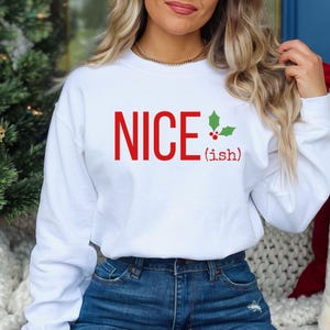 May include: White sweatshirt with the text "NICE (ish)" in red letters and a green holly sprig with red berries.