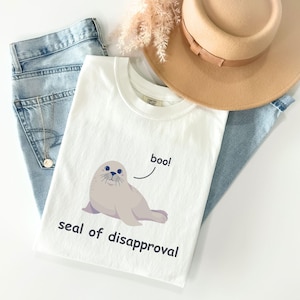 Peut inclure: T-shirt blanc avec un dessin de phoque de dessin animé disant "boo !" et "seal of disapproval". Le t-shirt est posé sur un jean bleu clair, un chapeau beige et un collier doré.