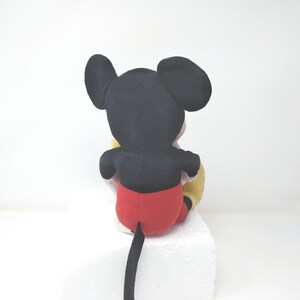 May include: A plush Mickey Mouse toy, viewed from the back, with black ears, a black body, red shorts, and a long black tail. The toy is sitting on a white surface, with a yellow glove visible.