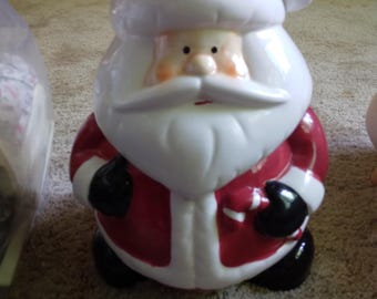 Puede incluir: Una figura de cerámica de Santa Claus con un traje rojo brillante con ribetes blancos, un gorro rojo a juego con un pompón blanco, una barba blanca, guantes negros y botas negras. La figura sostiene un bastón de caramelo y tiene un rostro pintado.