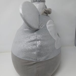 May include: A plush, gray elephant-shaped pillow with large, sparkly ears and a trunk. The pillow has a two-tone design, with a shimmering silver top and a solid gray bottom. The elephant's face is detailed with white accents.