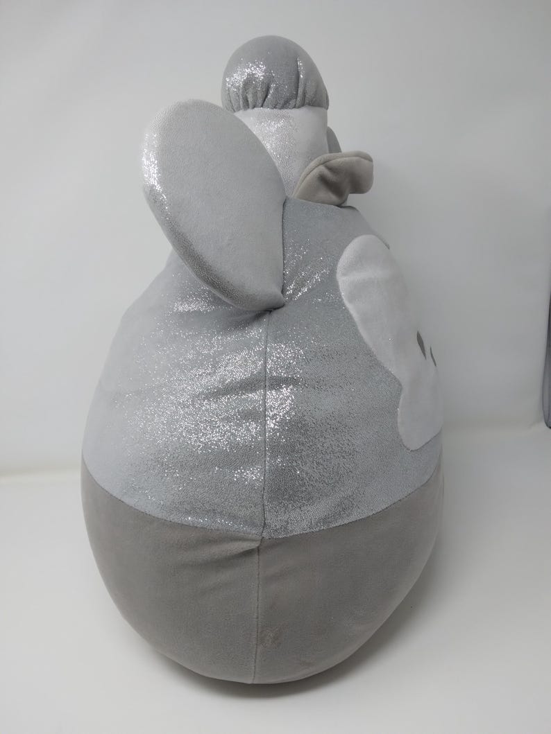 May include: A plush, gray elephant-shaped pillow with large, sparkly ears and a trunk. The pillow has a two-tone design, with a shimmering silver top and a solid gray bottom. The elephant's face is detailed with white accents.
