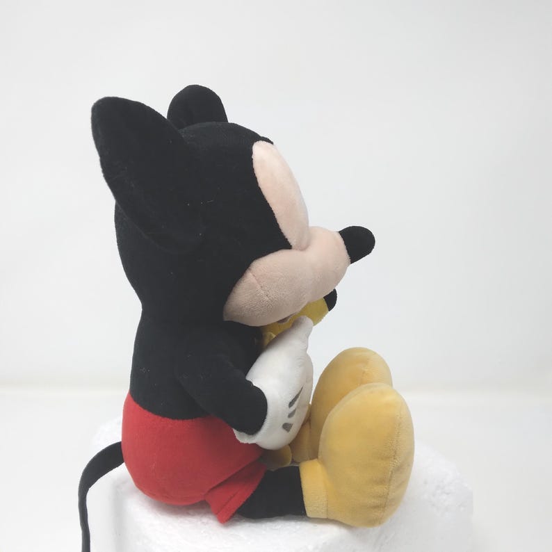 May include: A plush Mickey Mouse toy, viewed from the side, with black ears and head, a tan face, white gloves, red shorts, and yellow shoes. The classic Disney character is seated, with a black tail visible.