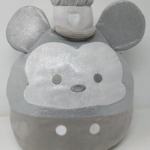 May include: A plush, round, gray character with large ears and a chef's hat. The character's face is white with gray eyes and a gray nose. The hat and ears have a glittery finish, and the bottom portion is a darker gray.
