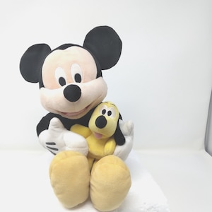 May include: A plush Mickey Mouse doll in black, white, and tan, holding a yellow Pluto doll. Mickey has large black ears, a white face, and a black nose. Pluto has black ears and a pink tongue. The dolls are on a white background.