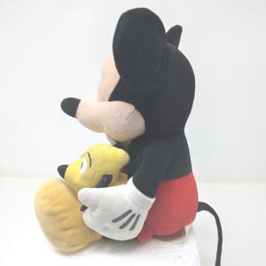 May include: A plush Mickey Mouse doll with black ears, a black head, and a red bottom. Mickey is holding a yellow plush Pluto doll. Mickey has white gloves with three black lines on each hand. The dolls are on a white surface.