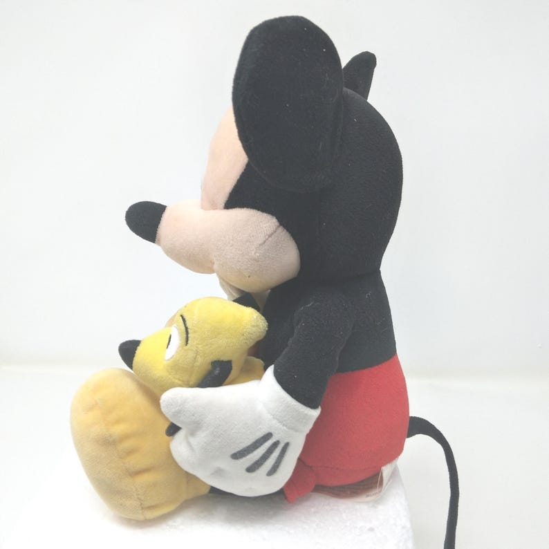 May include: A plush Mickey Mouse doll with black ears, a black head, and a red bottom. Mickey is holding a yellow plush Pluto doll. Mickey has white gloves with three black lines on each hand. The dolls are on a white surface.