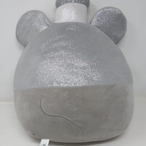 May include: A plush, gray elephant-shaped pillow with sparkly silver accents on the ears and top of the head. The pillow has a rounded body and a small, embroidered smile. A white tag with the Disney logo is attached.