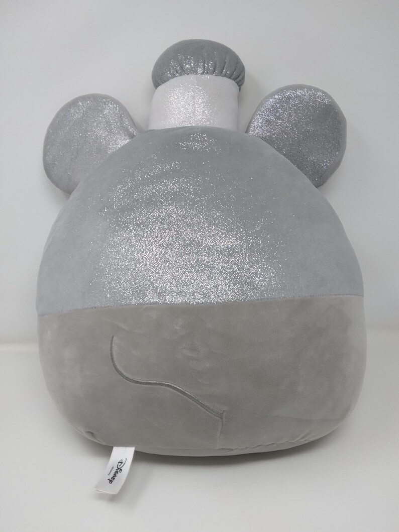 May include: A plush, gray elephant-shaped pillow with sparkly silver accents on the ears and top of the head. The pillow has a rounded body and a small, embroidered smile. A white tag with the Disney logo is attached.