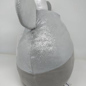 May include: A plush toy in the shape of an elephant, primarily gray with silver glitter accents. The elephant has large, soft ears and a rounded body. A small tag with the word "Disney" is attached.