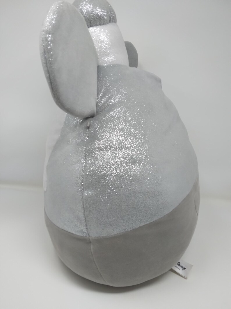 May include: A plush toy in the shape of an elephant, primarily gray with silver glitter accents. The elephant has large, soft ears and a rounded body. A small tag with the word "Disney" is attached.
