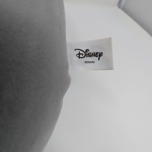 May include: A white rectangular tag with the Disney logo in black script. The tag is against a white background, with a gray object partially visible on the left side.