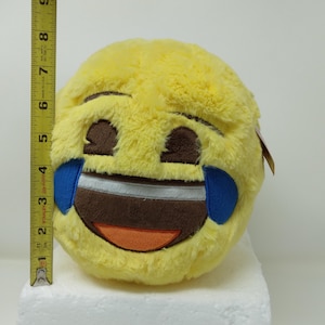 May include: A plush, round emoji pillow in bright yellow, featuring a laughing face with tears. The emoji has brown eyes and mouth, blue tears, and an orange tongue. The pillow measures approximately 8 inches in diameter.
