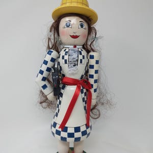 May include: A wooden doll with a yellow hat, brown hair, and a checkered blue and white dress. The doll has red shoes and a red ribbon tied around the waist. A black dog figurine is held in the doll's hand. The doll is standing on a round base.