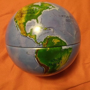 May include: A globe with a detailed map of the world, featuring continents in green and blue oceans. The globe is split in half, showing North and South America, with text labels for countries and oceans. The globe sits on an orange surface.