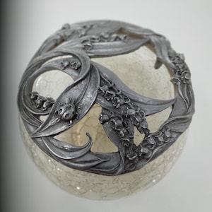 May include: A decorative glass orb with a crackled surface, topped with a silver-colored metal floral design. The metalwork features lily of the valley flowers and leaves, with a small ladybug accent. The orb is a unique home decor item.