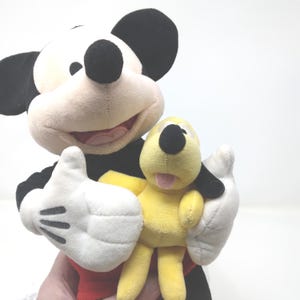 May include: A plush Mickey Mouse doll with black ears, a black body, and white gloves holds a yellow Pluto doll. Mickey Mouse has a smiling expression with a red mouth. Pluto has a black nose and ears, and a pink tongue.