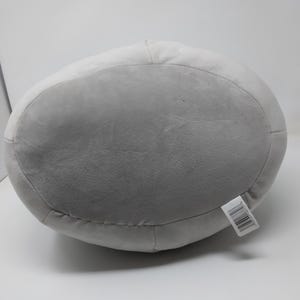 May include: A large, gray plush pillow with a rounded shape. The pillow has a lighter gray top and a darker gray bottom. A white tag with black text is attached to the side. The pillow is made of a soft, plush material.