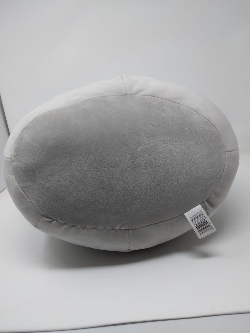 May include: A large, gray plush pillow with a rounded shape. The pillow has a lighter gray top and a darker gray bottom. A white tag with black text is attached to the side. The pillow is made of a soft, plush material.
