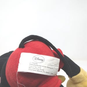 May include: A plush toy featuring a red body, black accents, and a yellow hand. A black handle is attached to the top. A white tag with the Disney logo and text is visible.